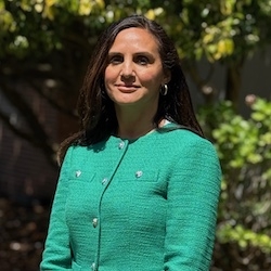 Sukhie Deep Kaur Bal - profile photo of woman with medium length dark hair wearing green suit jacket, with trees in background 